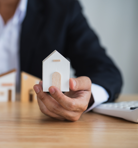 Makelaar in Malden houdt een wit houten huisje vast aan een bureau, symbool voor persoonlijke begeleiding bij verkoop, aankoop en taxatie van je woning.